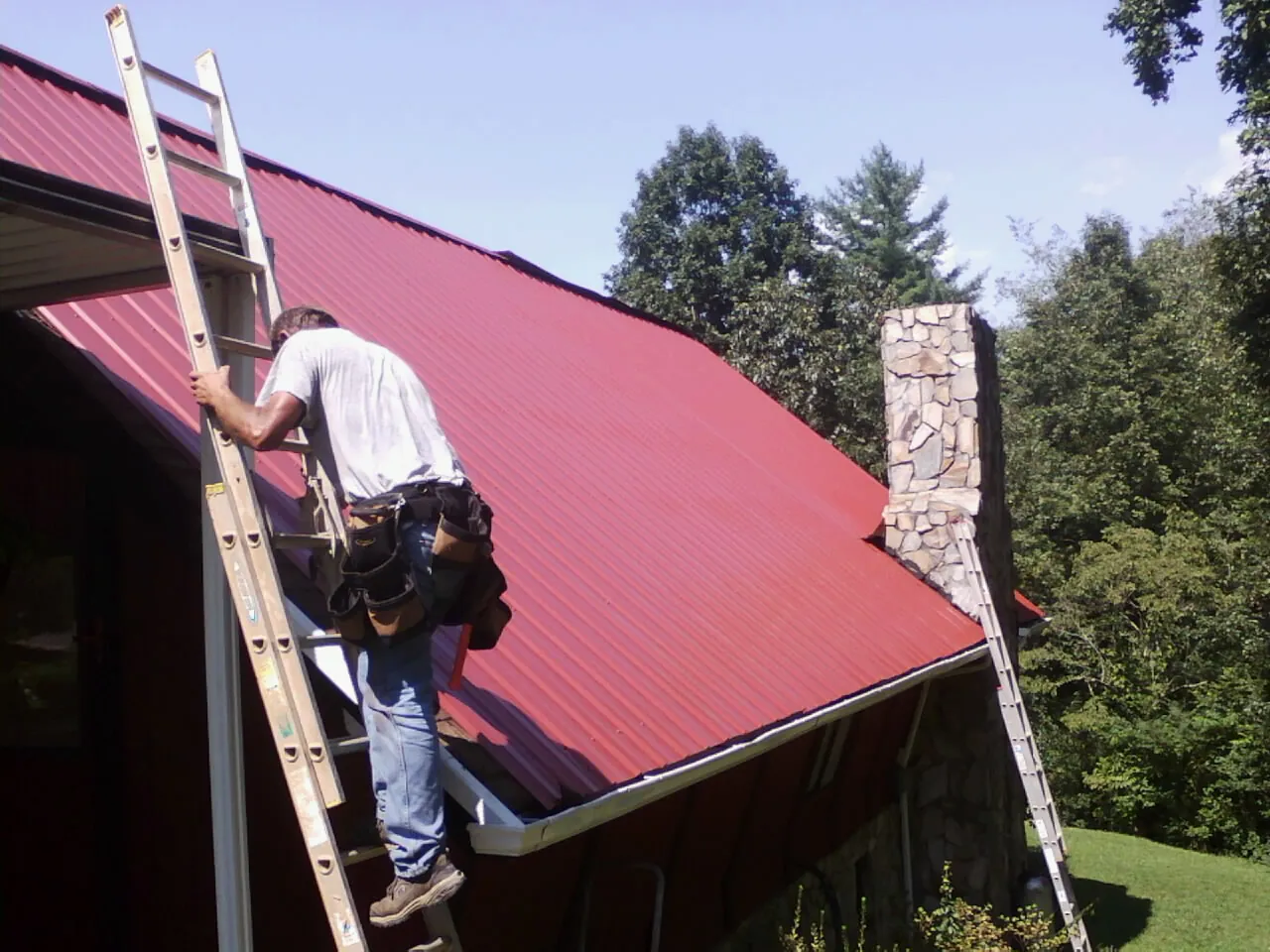 Roofer on a ladder beside a red metal roof and stone chimney — Soffit & Fascia Repair by Roofing Brothers in Turtletown
