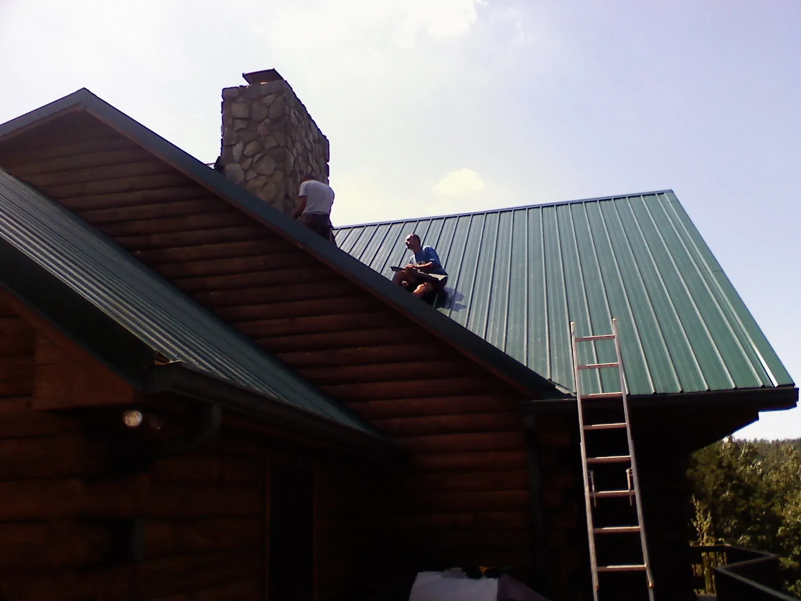 Roofers installing green metal panels beside a stone chimney on a log home in Turtletown